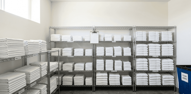 A bright, clean storage room with perfectly folded white towels and sheets stacked on wire shelving. This visual reinforces the T-200 linen and 3-par system.